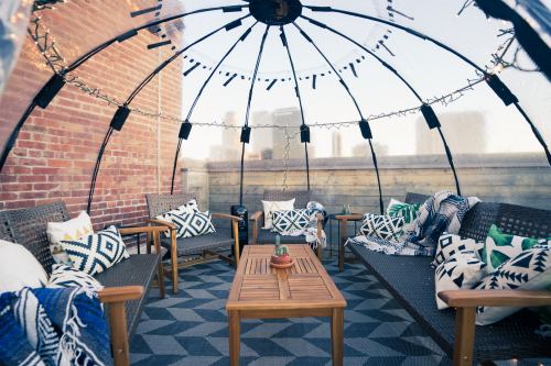 Cozy outdoor seating area in a dome, featuring patterned cushions, blankets, and string lights against a city skyline.