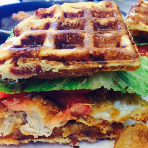 A close-up of a waffle sandwich filled with fried chicken, lettuce, and tomato, served with a side of chips.