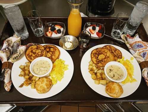 Two plates of breakfast with pancakes, scrambled eggs, potatoes, biscuits, and fruit, served on a wooden tray.