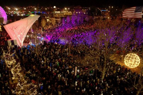 A large crowd gathers at night, illuminated by colorful lights and decorations, with trees and an American flag in the background.