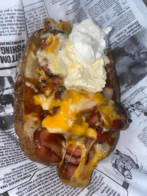 A loaded baked potato topped with cheese, hot dogs, and a dollop of sour cream, served on newspaper.