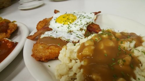 A plate of fried chicken with a sunny-side-up egg, bacon, and mashed potatoes topped with gravy, garnished with parsley.