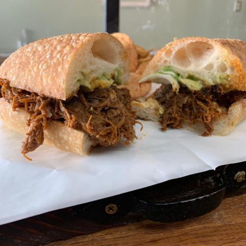 A close-up of a sandwich filled with shredded meat and avocado, served on a crusty roll, with a side of chips.