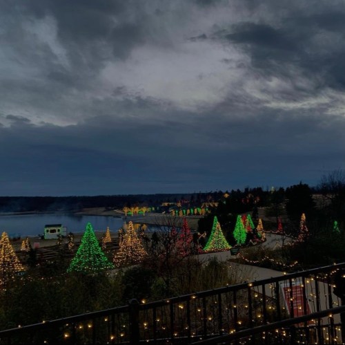 A scenic view of a lakeside area adorned with colorful, illuminated Christmas trees under a cloudy sky.
