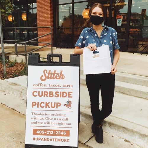 A masked employee stands by a curbside pickup sign for Stitch, holding a takeout bag and smiling.