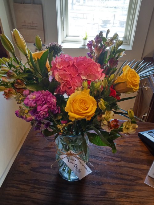 A vibrant bouquet of mixed flowers in a glass vase, featuring pink hydrangeas, yellow roses, and various greenery.