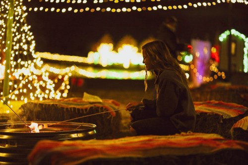 A person sits by a fire pit, roasting marshmallows, surrounded by festive lights and hay bales at night.