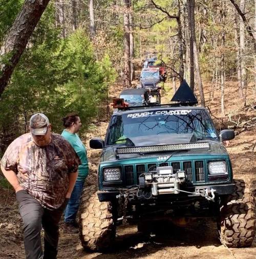 A group of off-road vehicles lined up on a forest trail, with a person walking nearby.