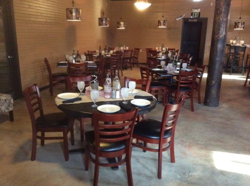 A cozy restaurant interior with wooden tables, chairs, and dim lighting, set for dining with glasses and plates.