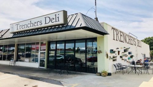 Exterior of Trencher's Deli featuring a sign, outdoor seating, and a sunny day.