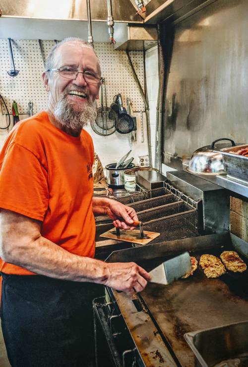A smiling man with a beard cooks on a grill in a kitchen, wearing an orange shirt and using a spatula.