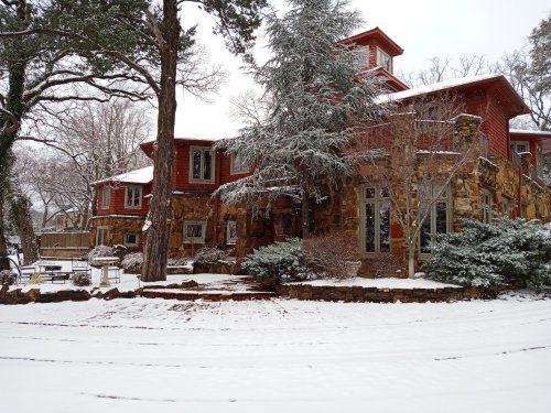 A snow-covered house with stone and wood exterior, surrounded by trees and a snowy yard.