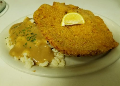 A plate of fried fish with a lemon slice, served alongside mashed potatoes topped with gravy and parsley.