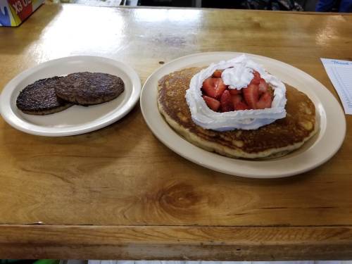 A plate with a large pancake topped with whipped cream and strawberries, alongside a plate of two sausage patties.