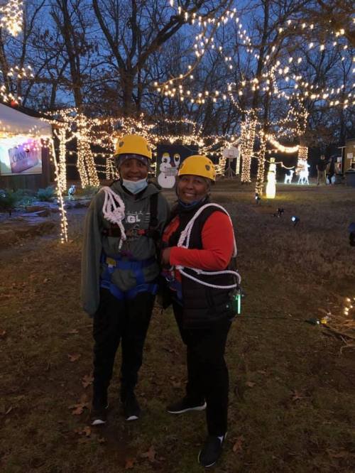 Two women wearing helmets and harnesses stand outdoors, smiling in front of a festive, light-decorated scene.