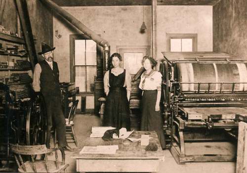 Three people stand in a vintage printing shop, surrounded by printing equipment and wooden furniture.