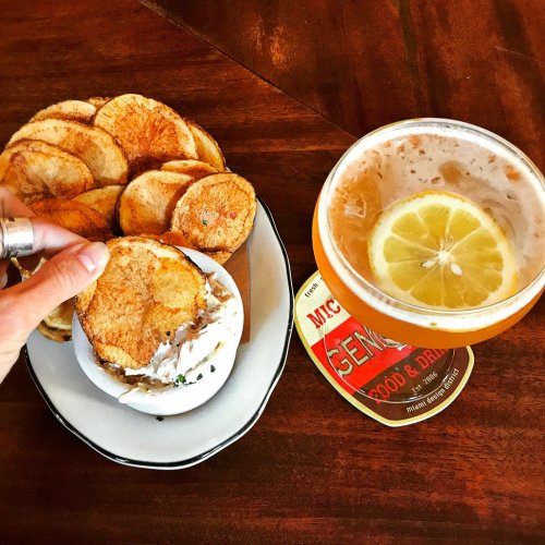 A hand holding a potato chip above a bowl of dip, with a glass of beer and a coaster on a wooden table.