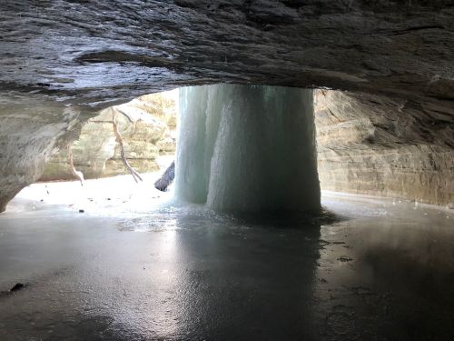 A frozen waterfall inside a rocky cave, with ice formations and a reflective icy surface.