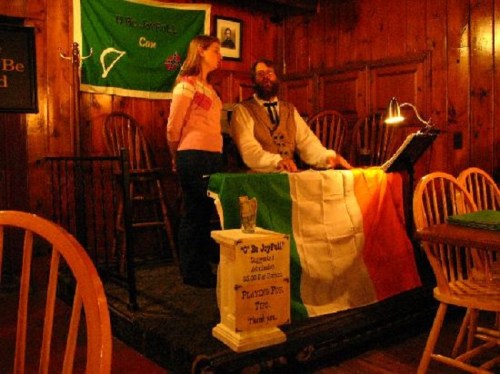 A man and woman stand at a table with an Irish flag, in a cozy wooden room with chairs and a sign on the wall.