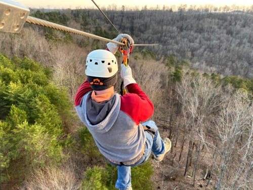 A person in a helmet zip-lining over a forested area during sunset, showcasing trees and a clear sky.