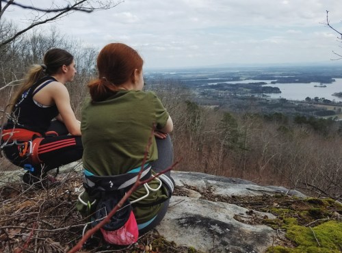 Two hikers sit on a rocky ledge, overlooking a scenic view of a lake and distant landscape.