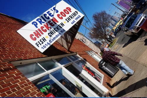 A person walks past Price's Chicken Coop, a restaurant sign visible on a brick building, with a clear blue sky above.