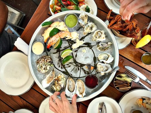 A seafood platter with oysters, shrimp, and sauces, surrounded by plates of salad and drinks on a wooden table.