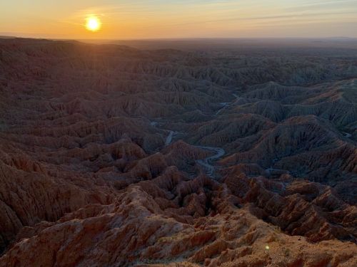 A stunning sunset over rugged, undulating desert terrain with deep canyons and valleys.