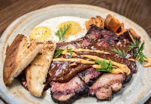 A plate of sliced brisket with barbecue sauce, two fried eggs, sweet potato fries, and toasted bread, garnished with herbs.