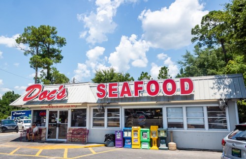 A seafood restaurant named "Doc's Seafood" with a bright sign, outdoor seating, and nearby newsstands under a blue sky.