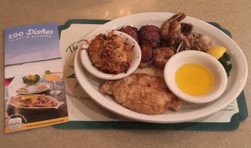 A plate of seafood including fish, shrimp, and crab cakes, served with a side of butter and a brochure about Alabama dishes.