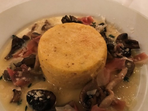 A round serving of polenta topped with sautéed mushrooms and prosciutto, drizzled with a creamy sauce.