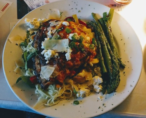 A plate of pasta topped with grilled chicken, vegetables, cheese, and asparagus, garnished with herbs.