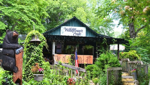 A charming café surrounded by greenery, featuring a bear statue and a sign that reads "Wildflower Café."
