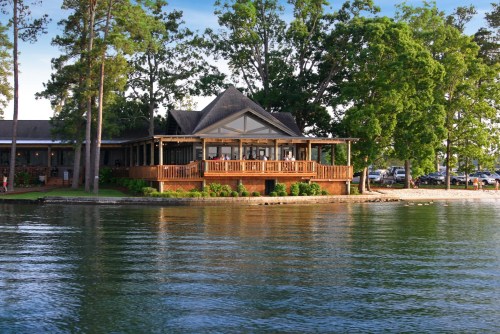 A lakeside view of a wooden building surrounded by trees, reflecting in the calm water.