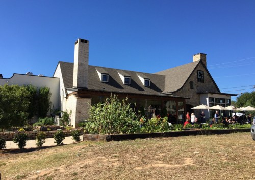 A charming stone building with a sloped roof, surrounded by greenery and outdoor seating under umbrellas on a sunny day.
