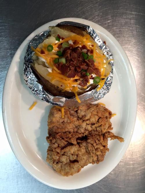 A baked potato topped with cheese, bacon, and green onions, served alongside a piece of fried chicken on a white plate.