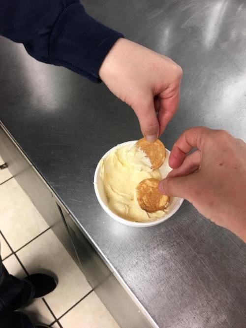 Two hands reaching into a bowl of ice cream, each holding a cookie. The setting is a kitchen with a stainless steel surface.
