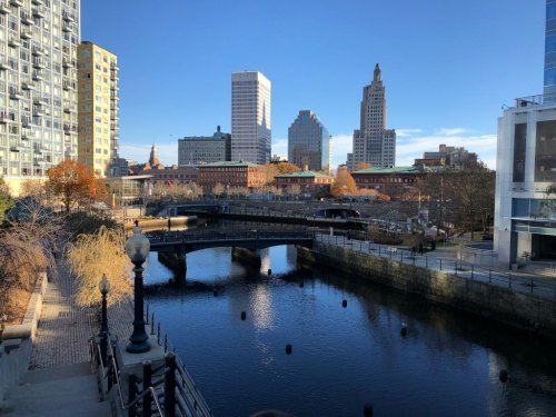 City skyline with modern buildings, a river, and a bridge, under a clear blue sky. Trees with autumn foliage in the foreground.