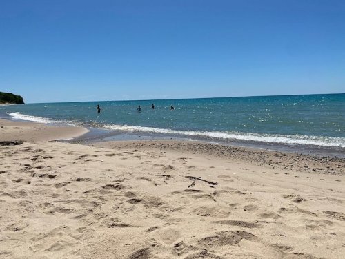 A sunny beach scene with people wading in the clear blue water and a sandy shore under a bright blue sky.