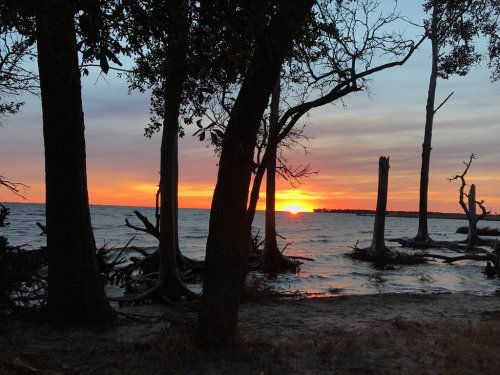 Sunset over a calm lake, framed by silhouetted trees and a sandy shore, with vibrant orange and purple hues in the sky.