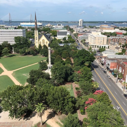Aerial view of a city park with trees, a monument, and buildings, with a bridge and water in the background.