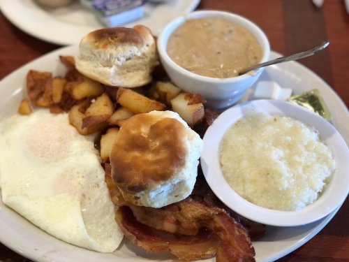 A hearty breakfast plate with eggs, biscuits, bacon, fried potatoes, grits, and a bowl of gravy.