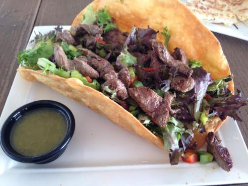 A large taco filled with grilled beef, fresh greens, and vegetables, served with a side of green salsa.