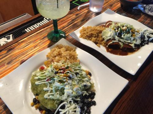 Two plates of Mexican food: one with enchiladas and rice, the other with green sauce, black beans, and a margarita.