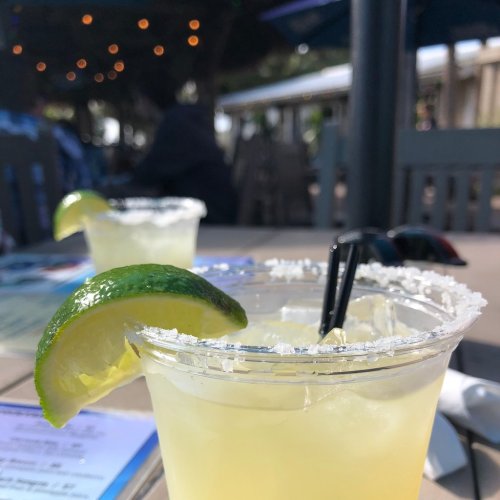 Two refreshing margaritas with lime wedges and salt rims, set on a table with a blurred background of outdoor seating.