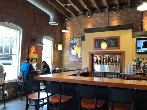 A cozy bar with a wooden counter, brick walls, and colorful posters, featuring a row of beer taps and patrons seated nearby.