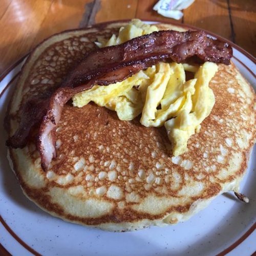 A fluffy pancake topped with scrambled eggs and crispy bacon on a plate.