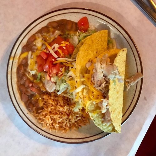 A plate with two tacos filled with chicken, lettuce, and cheese, served with rice and refried beans on the side.