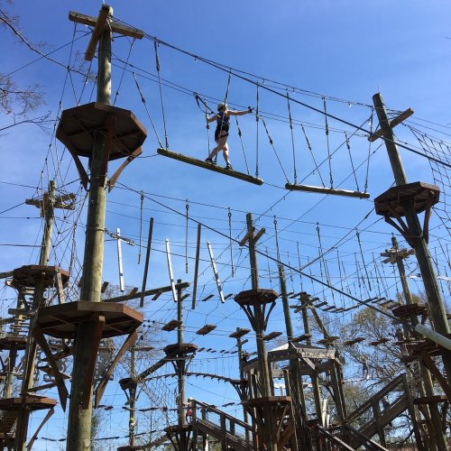 A person navigates a high ropes course with wooden beams and cables against a clear blue sky.